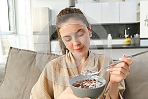 Image of smiling, happy young woman eating breakfast, holding bowl of cereals with milk, having meal at home