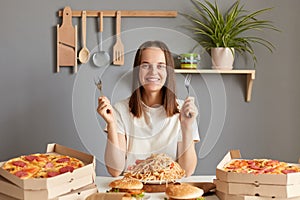 Image of smiling satisfied young adult woman with brown hair wearing white casual T-shirt sitting at table in kitchen, holding