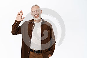 Image of smiling senior man waving hand, saying hello, greeting you, standing over white background