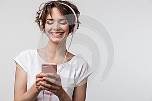 Image of smiling woman in basic t-shirt holding smartphone while listening to music with headphones