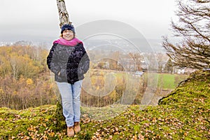 Image of a smiling woman with a black coat, denim jeans and a pink scarf resting on a trunk of a tree