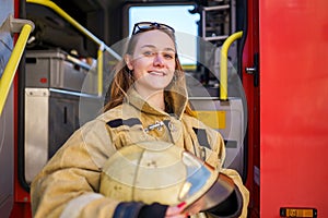 Image of smiling woman firefighter with glasses on head with helmet in hands standing next to fire truck