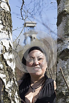 Image of a smiling woman in the middle of two tree trunks with a tower of a mine background