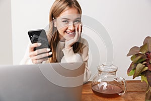 Image of smiling woman using laptop and smartphone while drinking tea