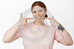 Image of smiling young redhead woman showing her white perfect smile, pointing at whitened teeth without braces