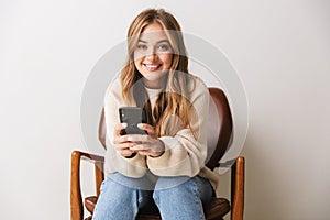 Image of smiling young woman using cellphone while sitting on armchair