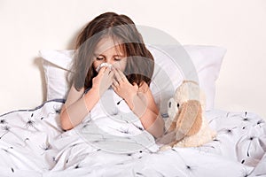 Image of sneezing sick little girl with fair hair, covering mouth with napkin, blowing nose, feeling unwell, getting ill, having