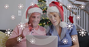 Image of snowflakes over senior caucasian couple wearing santa hats and using tablet