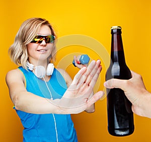 Image of sporty woman with dumbbell refuses from bottle of beer in studio.