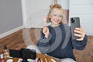 Image of stylish young woman, social media influencer, taking pictures on mobile phone, doing makeup tutorial for