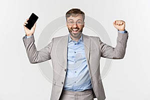 Image of successful businessman in grey suit and glasses, holding mobile phone, raising hands up and shouting yes from