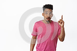 Image of surprised, curious african american man pointing finger up, lookin at top advertisement, showing sale