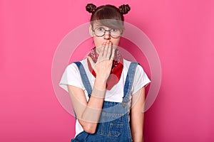 Image of surprised dark haired teenager with funny banches, covers mouth with hand, wears t shirt, overalls, glasses and bandana,