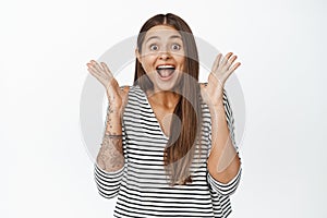 Image of surprised and happy woman reacts to big news, shaking hands near face and shouting from joy, white background