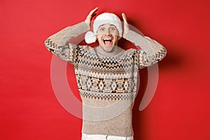 Image of surprised and happy young man in santa hat and christmas sweater, receiving amazing gift, standing excited