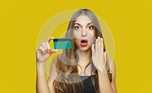 Image of surprised young lady standing over yellow background and holding debit card in hands. Looking at camera