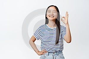 Image of teen asian girl winking and smiling, showing okay, ok gesture, assuring you, confirm something is good