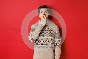 Image of tired or bored handsome man in winter sweater, yawning and covering mouth with hand, standing exhausted against