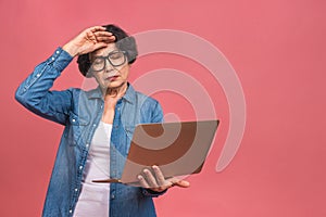 Image of tired depressed mature aged senior business woman standing isolated over pink background using laptop computer. Portrait