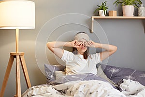 Image of tired sleepy woman with sleeping mask lying in bed and rubbing her eyes, being sleepless, having lack of sleep, wearing
