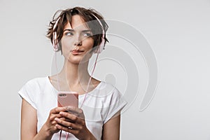 Image of tricky woman in basic t-shirt holding smartphone while listening to music with headphones