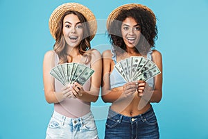 Image of two delighted girls 20s with different type of skin wearing straw hats rejoicing and holding fans of dollar banknotes, i