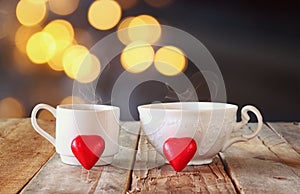 Image of two red heart shape chocolates and couple cups of coffee on wooden table in front of bokeh abstract background