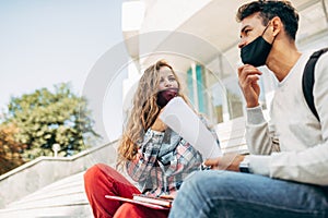 Image of two young students in protective face masks sitting on the stairs of the college campus outside and talking. A young