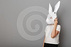 Image of unrecognizable woman wearing white t shirt and paper rabbit mask standing isolated over gray background, talking on smart