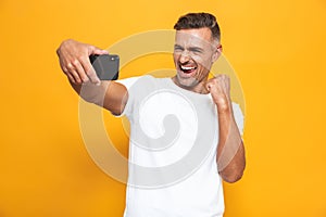 Image of unshaved man 30s in white t-shirt smiling and taking selfie photo on mobile phone