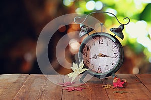 Image of vintage alarm clock next to autumn leaves on wooden table in front of abstract blurred background. retro filtered