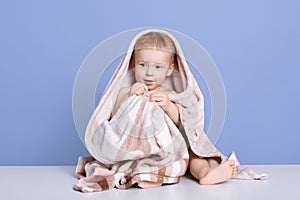 Image of wet baby boy wrapped in towel after washing him up, sitting against blue studio wall, looking aside, infant posing after