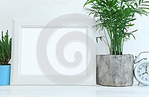 Image of white empty photo frame next to home plant over wooden table. For mockup, can be used for photography montage and text