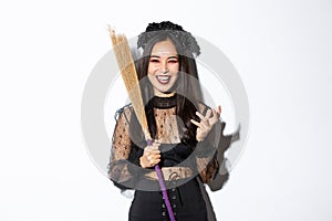 Image of witch in gothic lace dress and black wreath making evil laugh and holding broom, celebrating halloween