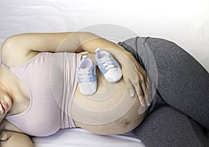 Image of A woman pregnant reclining on bed and touching or caressing her belly with baby shoes.