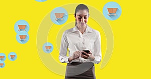 Image of woman using smartphone against multiple shopping cart icons on yellow background