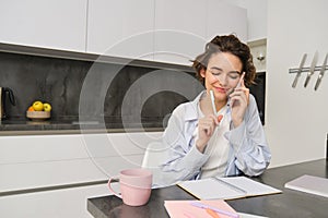 Image of woman working at home, making a phone call, sitting with smartphone, surrounded with paperwork, doing homework