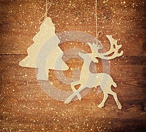 Image of wooden decorative christmas tree and reindeer hanging on a rope over wooden background with glitter overlay.