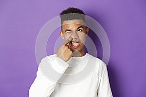 Image of young african-american man picking nose and looking upper left corner spaced out, standing over purple