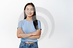 Image of young asian female model cross arms on chest, looking confident and smiling at camera, standing in casual