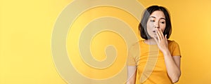 Image of young asian girl student, yawning, feeling tired, standing sleepy agaisnt yellow background