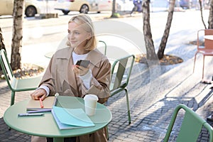 Image of young beautiful girl, student studying, doing homework in cafe, using mobile phone, holding smartphone, smiling