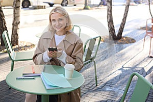 Image of young beautiful girl, student studying, doing homework in cafe, using mobile phone, holding smartphone, smiling