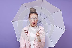 Image of young beautiful woman with hair bun holding umbrella, wears glasses, standing isolated over purple background, looks