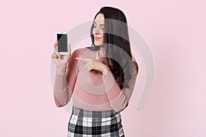 Image of young beautiful woman holding smart phone and showing at its blank screen isolated over pink background, female with long