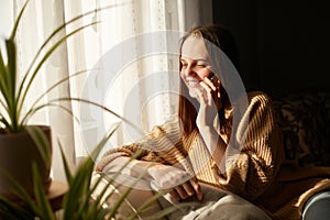 Image of young beautiful woman talking on smartphone with happy expression at home near window in sunlight, looking away with