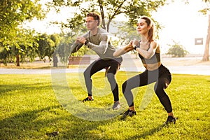 Image of young caucasian sporty man and woman 20s in tracksuits, doing workout and squatting together in green park during sunny