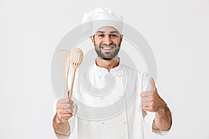 Image of young chief man in cook uniform smiling while holding wooden kitchen utensils