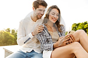 Image of young couple smiling while sitting outdoors with cellphone