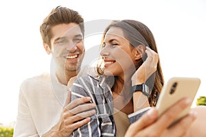 Image of young couple smiling while sitting outdoors with cellphone